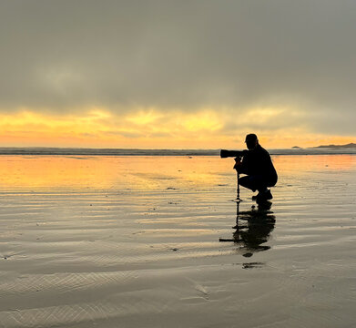 Silhouette of a man crouching on a beach taking a photo with a long lens camera on a monopod, British Columbia, Canada