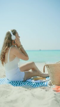 relaxed elegant woman on the ocean coast with straw bag and striped towel listening to the music with headphones.