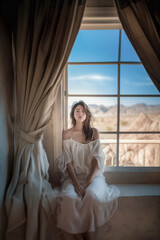 Obraz premium Woman in flowing white dress sitting by a window with a scenic desert view and soft sunlight.