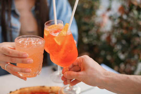 Close-up of three people clinking glasses with orange spritz and paloma cocktails at a pizzeria