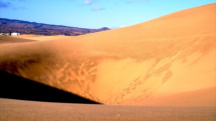 Wall of golden sand dunes with rocky mountains and blue sky in the background