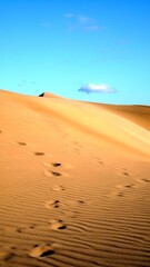 Footprints and texture on the golden sand wall of a desert dune with clear sky and isolated white clouds.