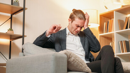 Fototapeta premium Headache pain. Health story. Business man suffering migraine on sofa in modern suit with lighted lamp in modern interior.