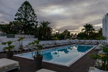 Empty swimming pool with sunbed in resort hotel. Vacation leisure. Blue water and reflection of palms in sunset. Pastel lounge chairs and close beach umbrellas. Tourism and advertising concept. Dusk