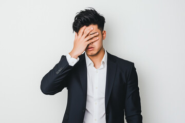 A young Japanese businessman in a suit covering his face with one hand, appearing frustrated or stressed, standing against a plain white background