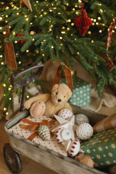 Close-up of a toy wagon with wrapped Christmas gifts, teddy bear and assorted ornaments next to an illuminated Christmas tree