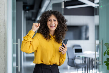 Young businesswoman enthusiastically celebrating success in office while holding phone. Bright...
