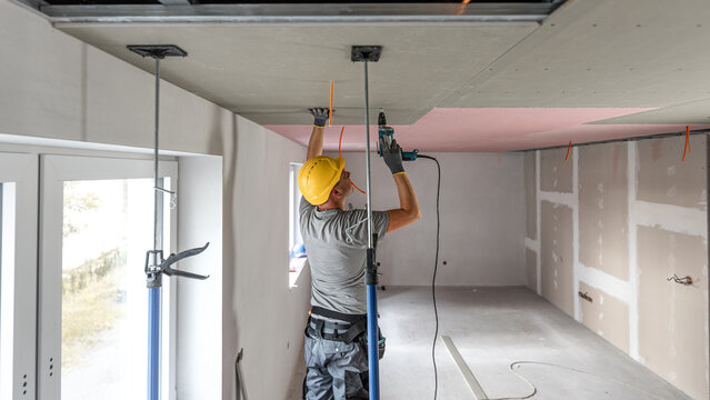 The worker screwing plasterboard to the ceiling. He is using special electric screwdriver and drywall lift. A red plasterboard improve the fire resistance of ceiling structures.