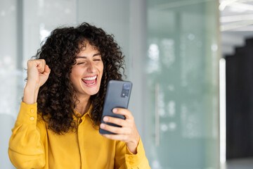 Joyful woman in yellow shirt celebrates success with smartphone in modern office setting....