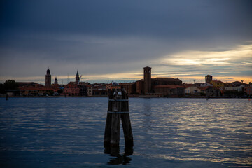 A wooden Venetian bricola (limit of the navigable canal)) in the Chioggia lagoon, in the blue of the sunset, with the houses in the background, Chioggia, Venice area, Italy