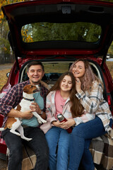 Happy parents with daughter and cute dog Jack Russel terrier sitting in car trunk on autumn day....