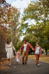 Fototapeta premium Happy family walking on green grass strewn with fallen leaves in Park in autumn. Parents, two children are walking in park at sunset with holding hands. Family, child, walk through Autumn Park.