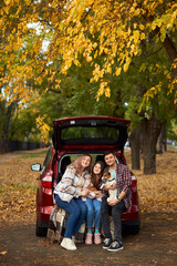 Happy parents with daughter and cute dog Jack Russel terrier sitting in car trunk on autumn day. Long auto journey break.