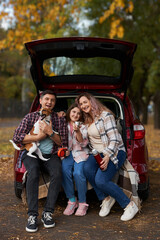 Happy parents with daughter and cute dog Jack Russel terrier sitting in car trunk on autumn day. Long auto journey break.