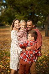Fototapeta premium Happy family walking on green grass strewn with fallen leaves in Park in autumn. Parents, two children are walking in park hug and laugh. Family, child, walk through Autumn Park.