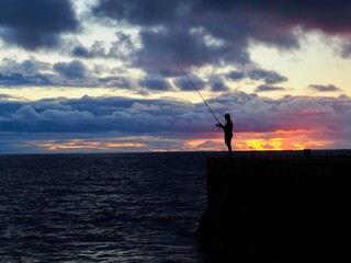 A lone person stands on a pier casting a fishing line into the sea as the sun sets, painting the sky with vibrant hues of orange, red, and purple. Peaceful and serene.