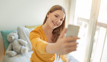 A teen girl with long hair smiles as she takes a selfie in a cozy bedroom. Soft sunlight filters in through the window, creating a cheerful atmosphere.