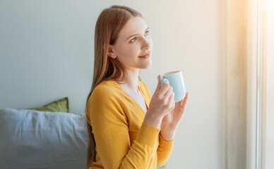 A girl with long hair, dressed in a yellow sweater, holds a light blue mug as she gazes thoughtfully out of a bright window. Sunlight creates a warm atmosphere in the room.