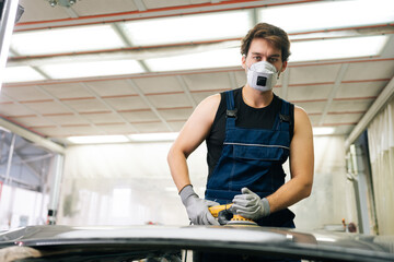 Portrait of skilled mechanic male in face mask polish hood using grinding unit in automobile repair and renew service station. Repair after an accident. Concept of car repair and maintenance work.