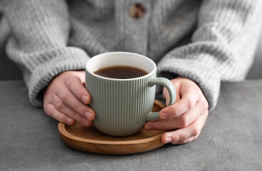 Woman in cozy sweater holding mug with espresso coffee close up on a gray table close up.