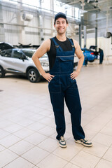 Full length vertical portrait of positive technician male in uniform and cap standing posing auto service center, smiling looking at camera, enjoying time at workplace. Concept of car maintenance