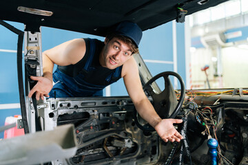Puzzled young mechanic gesturing inside disassembled car, showcasing automotive expertise and work in progress, looking at camera with puzzled emotion. Shooting in slow motion.