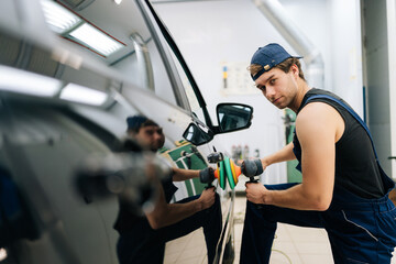 Portrait of confident mechanic male in uniform polish auto door using grinding unit in automobile repair and renew service station. Repair after accident. Concept of car repair and maintenance work.