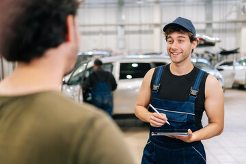 Positive mechanic male in uniform explaining checklist car maintenance and repair to unrecognizable man client at vehicle repair shop. Repairman with clipboard talking to man or owner at car shop.