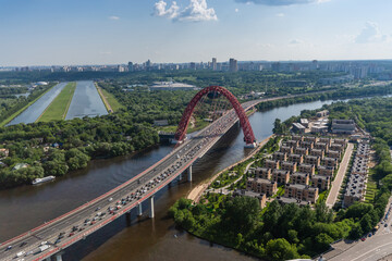 Fototapeta premium Aerial view of the red arch bridge spanning a river with urban landscape and greenery under a sunny sky