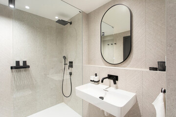 Modern design of bathroom in beige color, with white washbasin, oval mirror, white bathtub and black taps