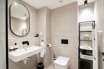 Bathroom in beige color with oval mirror on wall, white washbasin and toilet, black shelves for...