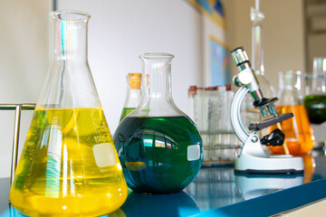 A close-up view of a laboratory setup featuring chemical solutions in various glassware, including Erlenmeyer flasks and round-bottom flasks  A microscope and a rack of test tubes 