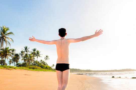 Young man wearing  a swimwear  is walking on the beach sand with his arms wide open, expressing well-being and gratitude during his beach trip. 