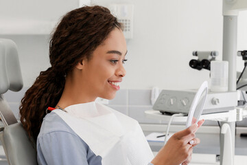 Fototapeta premium Beautiful black lady checking her white smile in mirror after stomatological treatment in modern clinic