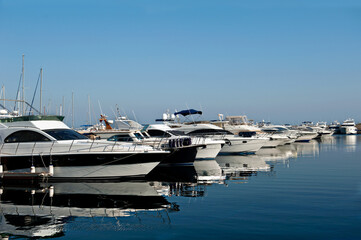 A lineup of luxurious yachts docked in a calm marina under a clear blue sky, their reflections mirrored perfectly in the tranquil water, capturing the elegance and relaxation of a coastal lifestyle.
