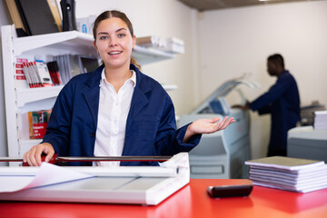 Portrait of cheerful young female typographer in uniform using paper cutter during work in printer...