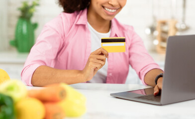 Ordering Food Online. Smiling black girl with laptop and credit card purchasing grocery delivery from internet in kitchen