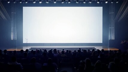 Audience members sit in a conference hall, attentively watching a blank white screen during a presentation event