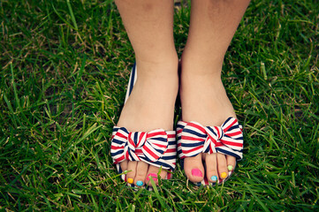 A close-up of a child's feet in colorful, striped sandals with large bows, standing on green grass. Each toenail is painted in a different bright color, creating a playful and cheerful scene.
