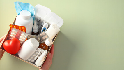 Medical supplies in a box in female hands: masks, pills, bottles, blister packs, and a thermometer. Medical donations, first aid kit, home delivery of medications concept.
