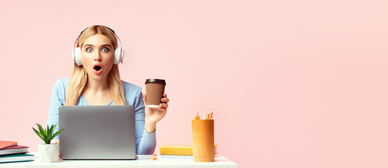 Deadline Concept. Shocked student in headphones sitting at desk and holding paper coffee cup over pink studio wall