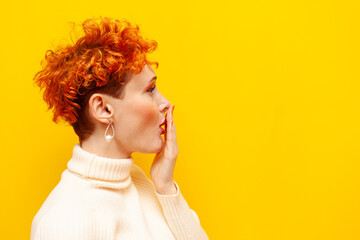 young red-haired excited woman with curly hair is surprised and looks away on yellow isolated...