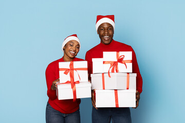 A couple dressed in bright red sweaters and Santa hats happily presents wrapped Christmas gifts with red ribbons, celebrating the festive holiday season together.