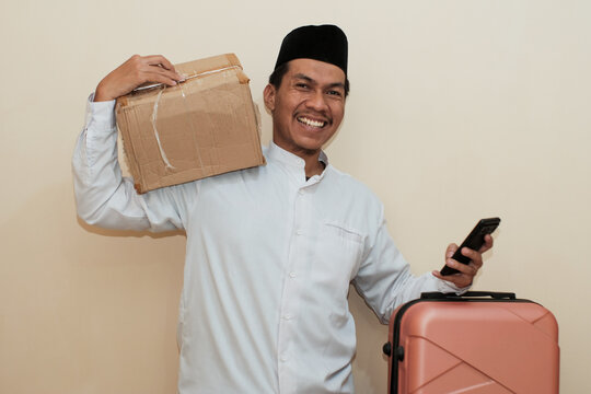 Smiling Southeast Asian muslim man carrying cardboard box and suitcase while holding mobile phone. Isolated on white background. Ideal for concepts of Ramadan and mudik travel.