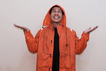 Smiling Southeast Asian man wearing orange raincoat standing with gesturing hand checking the...