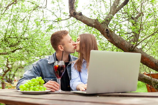 young couple sitting in garden at table drinking wine and using laptop on picnic in summer, woman and man kissing in park drinking alcohol and chatting online on computer