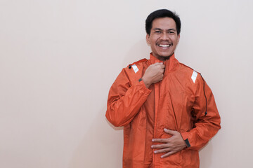 Smiling Southeast Asian man wearing orange raincoat with hands for zip up them looking at the camera. Positive person. standing over isolated white background. rainy season.