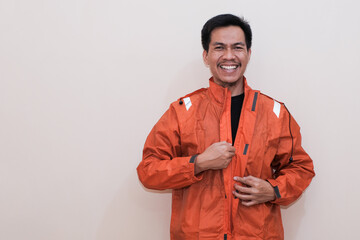 Smiling Southeast Asian man wearing orange raincoat with hands for zip up them looking at the camera. Positive person. standing over isolated white background. rainy season.