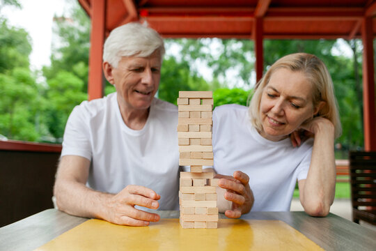 elderly couple of seniors in white t-shirts playing board game jenga in park outdoors, old man and woman building a tower of wooden bricks and competing in a game of balance