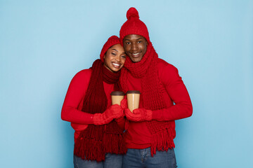 Fototapeta premium A couple dressed in coordinating red sweaters, hats, and gloves smiles while holding warm coffee cups. The bright blue background highlights their cheerful winter attire and expressions.